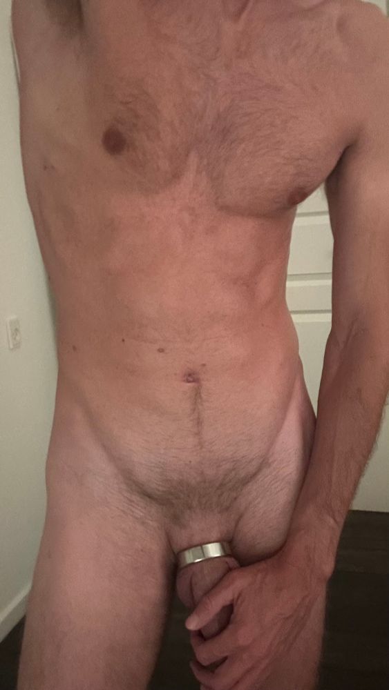 A gay man standing fully nude indoors with a metal cock ring around his hard dick and balls, gripping himself confidently. His muscular, hairy torso and dim lighting enhance the dominant, sexually charged energy as he invites the viewer to join in.