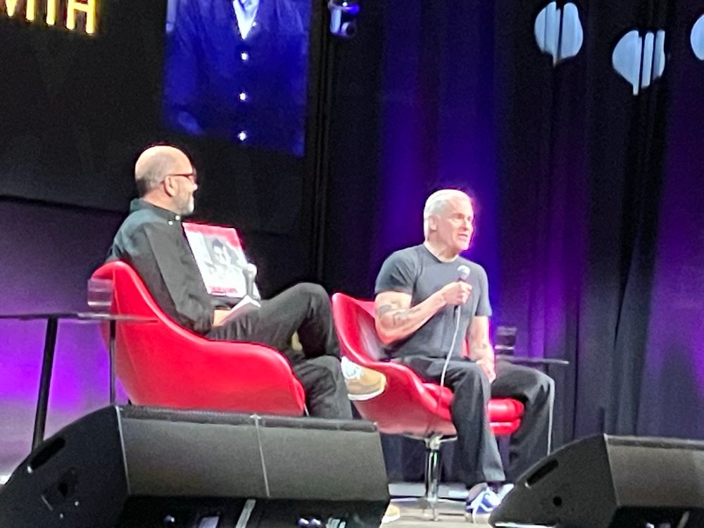 Henry Rollins and Nabil Ayers sitting in red chairs holding microphones in front of a 
purple backdrop.