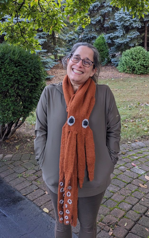 A smiling woman with her head cocked to the side stands in front of a brick path, with a lawn and trees in the background. She's wearing a khaki jacket and pants, and an orange knitted scarf in the shape of a KRAKEN with large eyes and white suckers on the backs of the tentacles