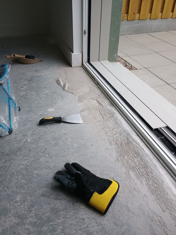 scene: new build floated concrete ground with workman glove in foreground, scraper in mid ground, dustpan with brush in background, open patio door to right, plastic-wrapped camping stool to left. out of frame, me covered in dust because fixing the mess builder leaves behind when flooring isn't included