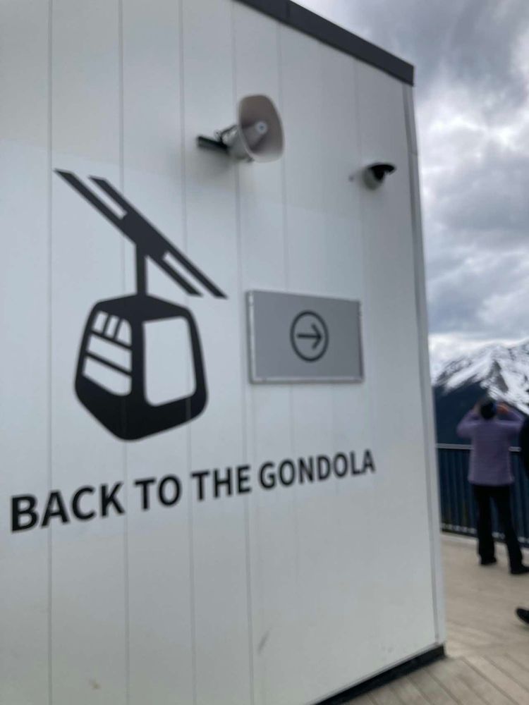 White panelled exterior wall with a graphic of a black gondola cage next to a black arrow pointing left with text below "Back to the Gondola', to the right is a person facing away and toward the snowy peaks of the Rocky Mountain, standing on the Banff observation deck.