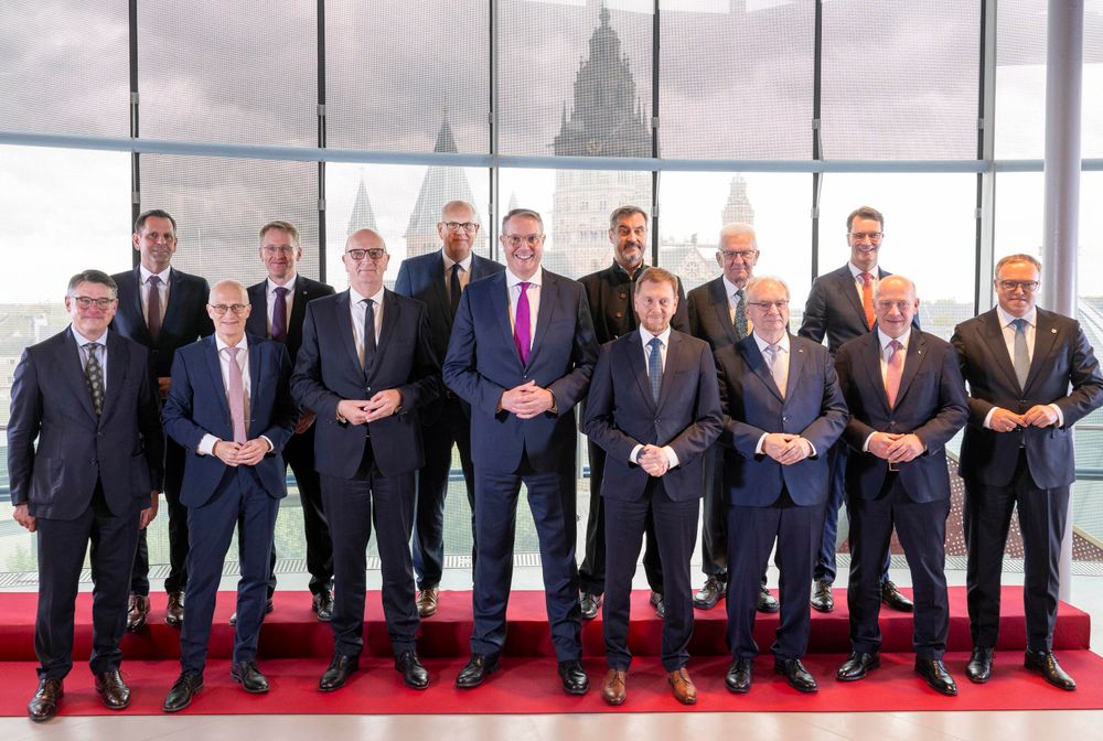 Eine Gruppe von Männern in zwei Reihen auf einem roten Teppich vor einer Glasscheibe, im Hintergrund ein religiöses Gebäude. Sie sind ausschließlich in dunklen Anzügen gekleidet, lediglich einer trägt keine Krawatte, es gibt erkennbar wenig Altersunterschiede. 