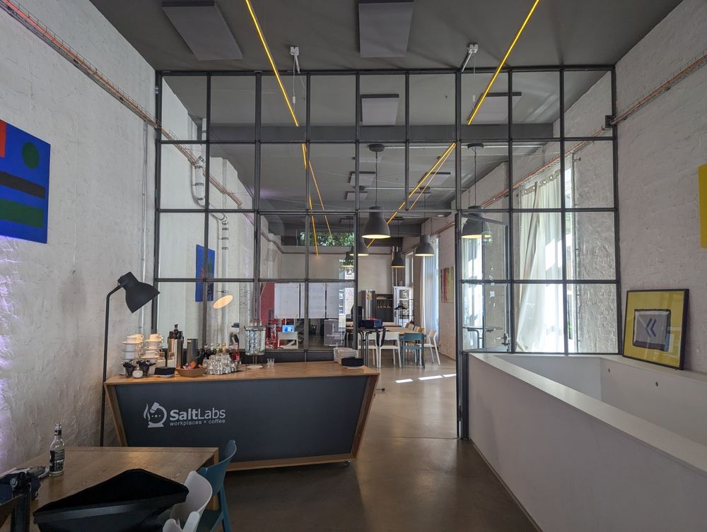 Ein Bereich eines Workshops in einem alten Industriegebäude. Weiße Wände, in der Mitte eine Wand aus Glas, das mit Metall eingegittert wurde. Darin eine Tür und dahinter ist ein großer Besprechungsraum zu erkennen. Davor ein Bar-Tisch mit der Aufschrift SaltLabs und Getränken darauf.