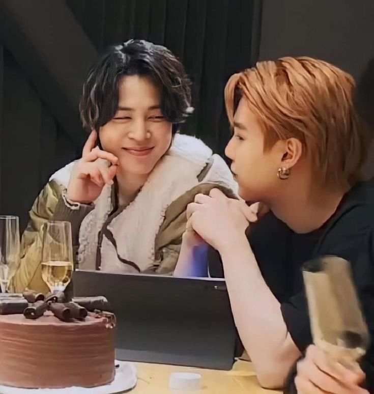 Jimin with dark hair and gray extensions (I love this hair on him) in a comfy beige-ish jacket with white sherpy comfy cozy trim, staring fondly at Yoongi who is wearing a black t-shirt, hands clasped in front of him. There is a computer on the table, and a chocolate cake.  Yoongi is not looking at the camera, saying something to Jimin.  I adore Yoongi's profile here.  