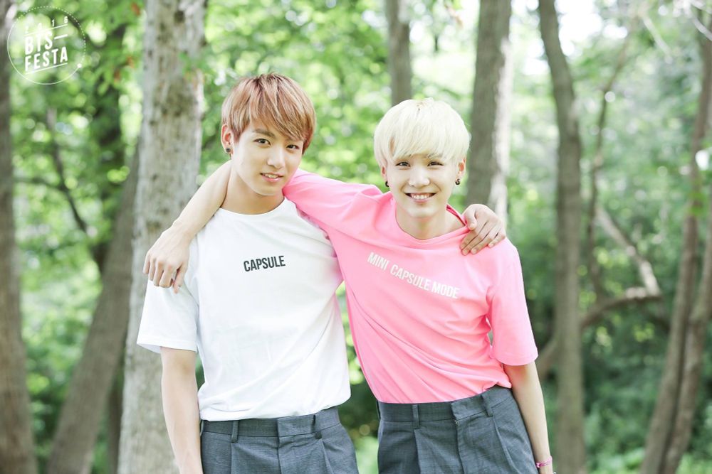 An outside shoot, Jungkook with Yoongi - Yoongi is wearing a pink "mini capsule mode" shirt and Jungkook a white "capsule" mode and boy was there speculation as to what those shirts were trying to say (hee hee).  Yoongi's hair is ghostly white, Jungkook's is light brown kissed by the sun. Both are wearing gray trouser-shorts, Yoongi's arm slung over Jungkook's neck, and really, this pic is so cute!  Trees behind them. 