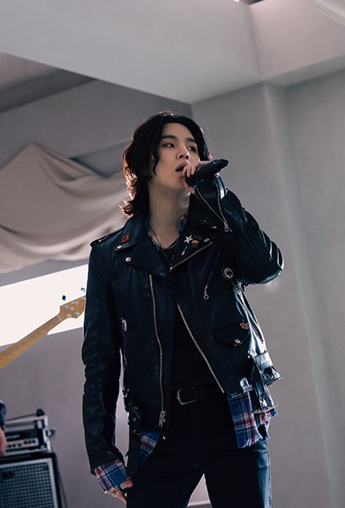 Yoongi in jeans, leather jacket over blue and red and white plaid shirt, in studio, long black hair, singing into his mic.  Iconic.