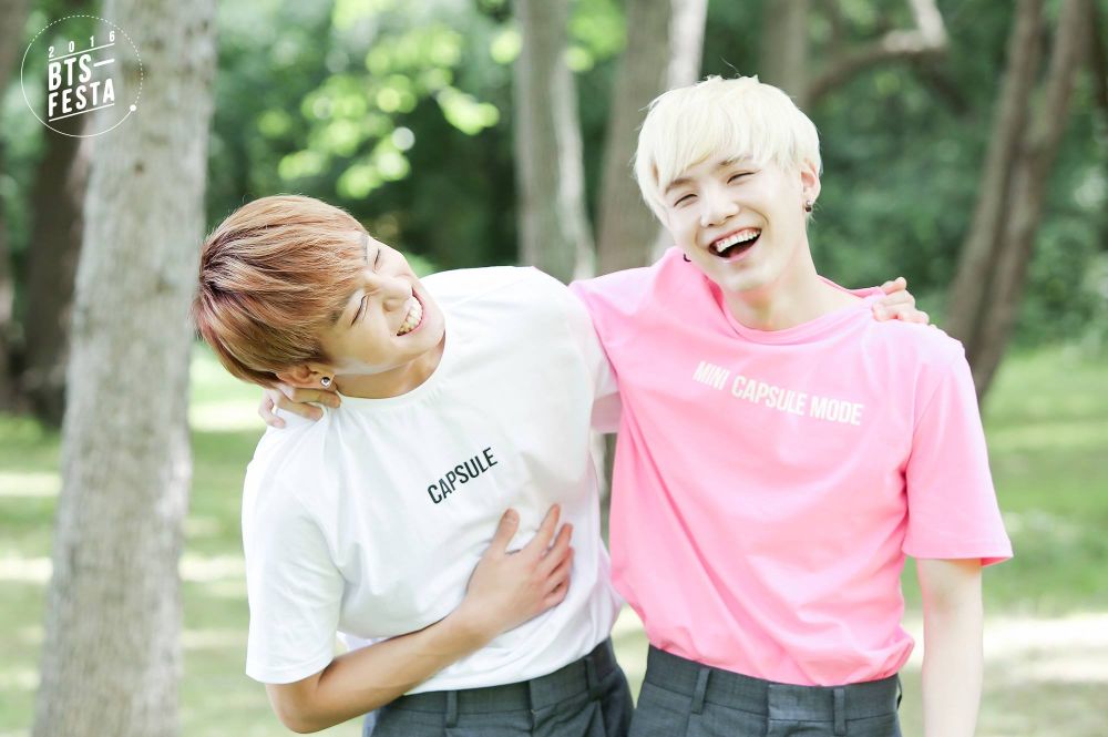 An outside shoot, Jungkook with Yoongi - Yoongi is wearing a pink "mini capsule mode" shirt and Jungkook a white "capsule" mode and boy was there speculation as to what those shirts were trying to say (hee hee).  Yoongi's hair is ghostly white, Jungkook's is light brown kissed by the sun. Both are wearing gray trouser-shorts. They have their arms around each other and are laughing.  Such a happy pic. 