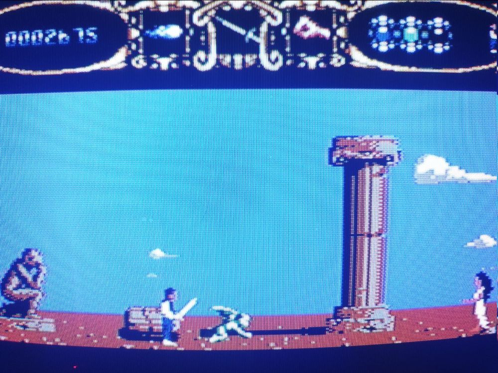 A gameplay snap of the second level in ancient Greece, with marble pillars already crumbling and falling over and a statue of a figure kneeling in thought. A lady in a toga and with black hair beckons to the protag.