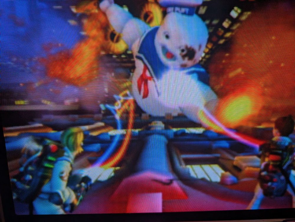 A snap of a dramatic battle against the Stay Puft Marshmallow Man burning up with two Ghostbusters seen from behind with Proton packs steaming at it 