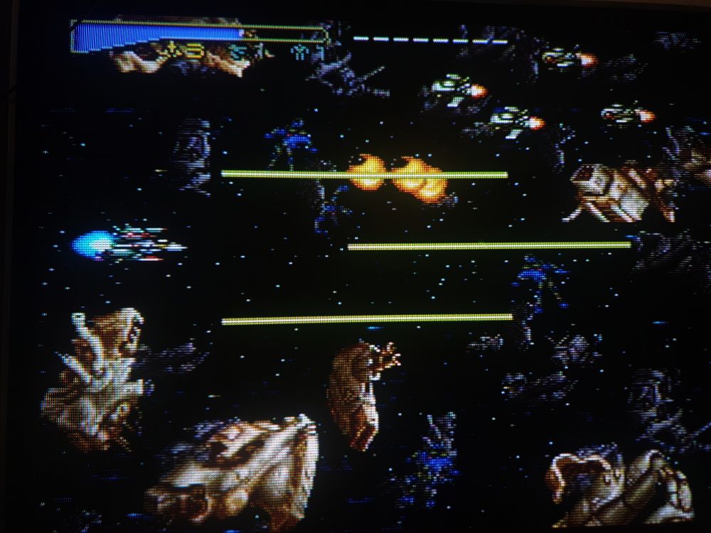 A crt screenie photo of the opening asteroid belt stage with the protag ship unleashing tri beams of later death as a squadron of enemy ships fly in 