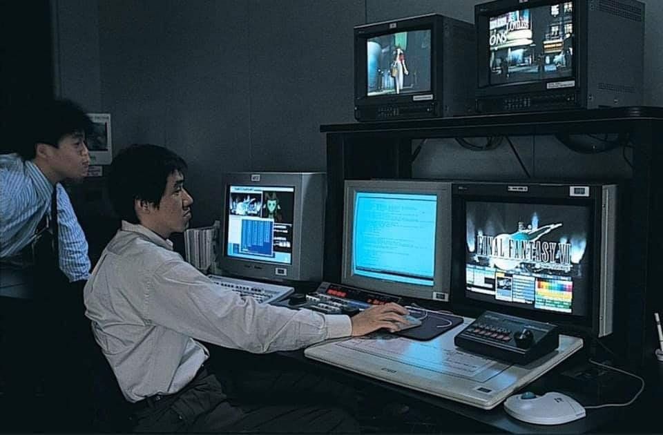 Two designers wearing suit shirts and ties sat and stood behind an array of five monitors of various sizes, two of them small PVMs on a shelf, along with keyboards and other computer tools. Various development screens for Final Fantasy VII can be seen.