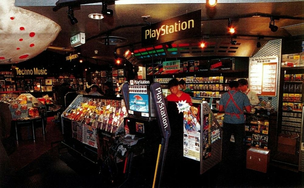 A snap of a photo of a scanned issue of Japan's The PlayStation Magazine with shelves and displays of a Dual Union store with a PS demo unit with Air Combat on the screen, PlayStation overhanging banners, and other games surrounded by shelves of music. It's quite moodily lit. 