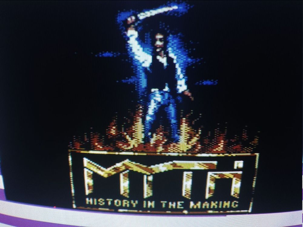 A close-up of the loading screen with the protag, a college kid who gets transported through time, holding up a sword and standing amid flame. 
