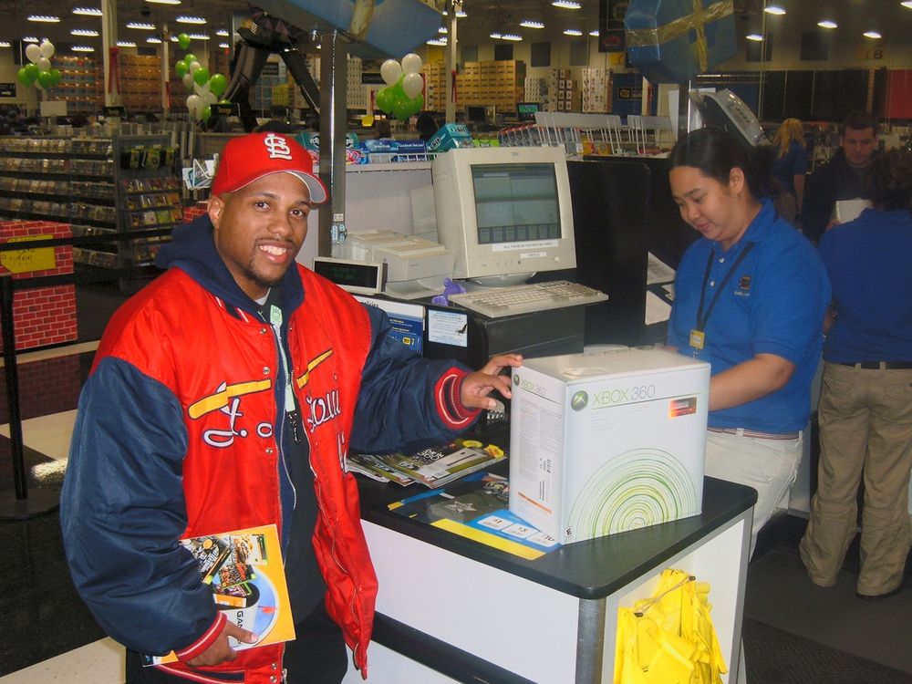 A black dude in a red hat and jacket smiling for the camera at a checkout with an Xbox 360 console boxed and ready to go.