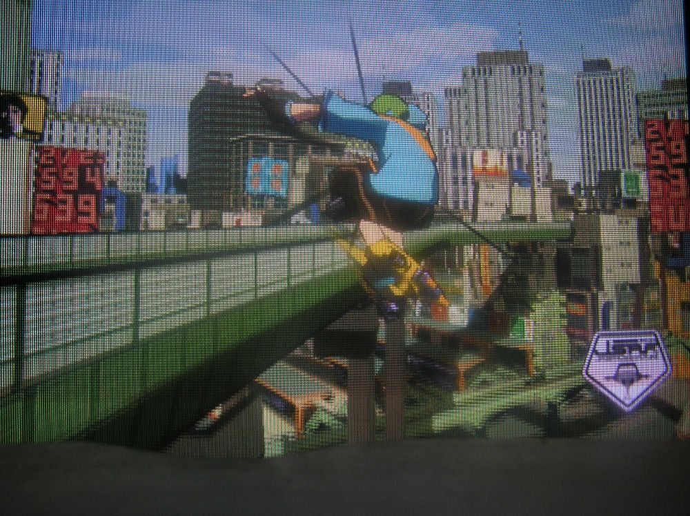 An action shot of another skater in a blue top and with yellow skates sailing through the air against a backdrop of a metropolis and highway