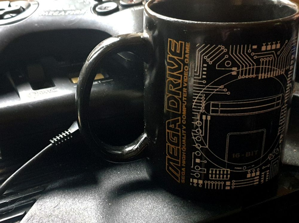 A photo of a Mega Drive themed black and gold mug next to Mega Drive mark I and Megafire mug 