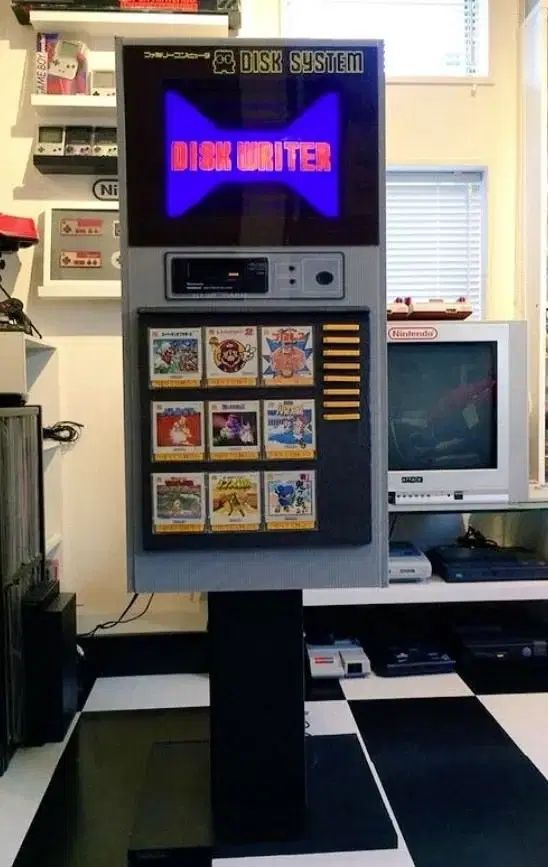 A shot of the colourful disk writer kiosk, a tall rectangle with a screen above a selection of game artwork squares in a 3x3 grid. Yellow FDS disks are stacked vertically to the right of them
