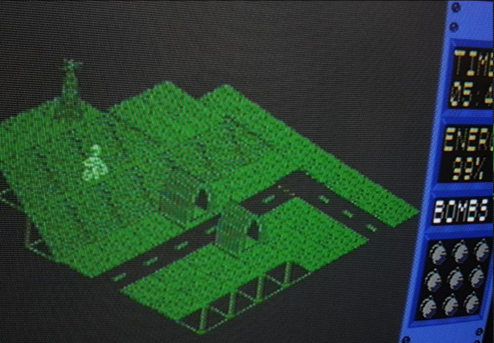A CRT TV photo of Glider Rider's pixelly isometric island world made up of flick screens to form a whole map. There are sloping hills, a tower, military facilities, and the player's bike. You have to flip the controls as you're going down a hill to transform and launch in your glider form and then drop bombs of specific facilities.