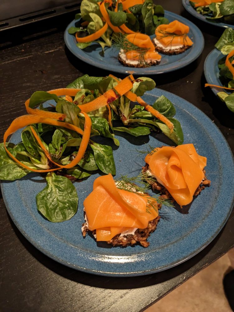 Auf einem blauen Steingut-Teller ist Feldsalat mit einigen Streifen Karotten drapiert, daneben sind zwei Frischkäse-Lachs-Tartes; im Hintergrund erkennt man weitere Teller mit gleichem Inhalt
