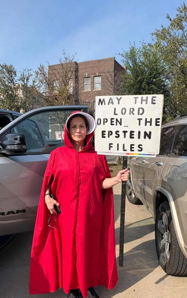 a woman dressed in red from the show The Handmaid's Tale and holding sign "may the lord open...the epstein files'