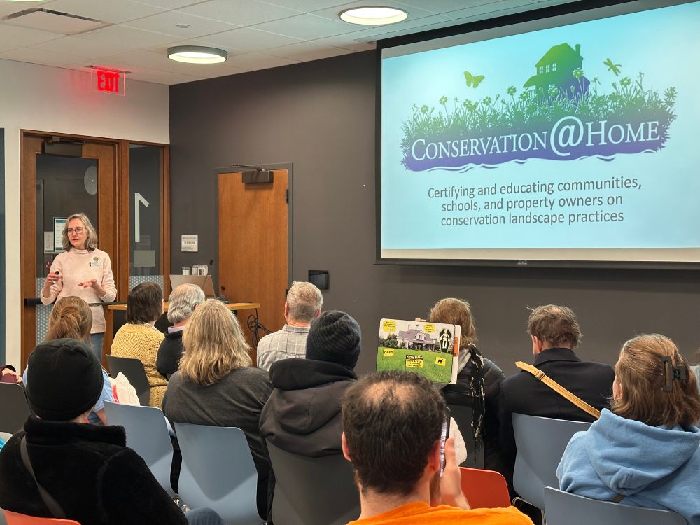 An Illinois Master Gardener speaking to a group of 30 people seated in chairs. The projector screen reads Conservation@Home Certifying and educating communities, schools, and property owners on conservation landscape practices 