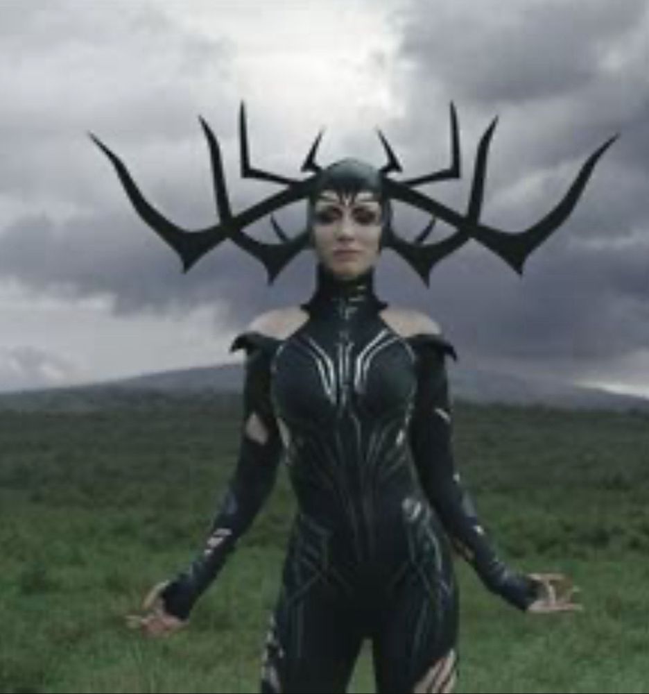 Hela from the movie Thor Ragnarok stands in a green field. She wears all black leather and a crown of 8 horns like upside down antlers.