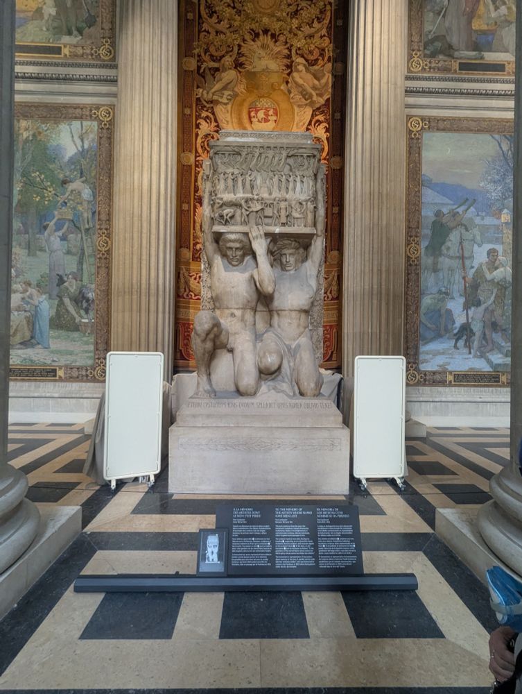 This monument is dedicated "TO THE MEMORY OF THE ARTISTS WHOSE NAMES HAVE BEEN LOST". 

• The work, created by Paul Landowski between 1912 and 1914, honors ordinary, unknown individuals rather than revolutionary figures. 
• It features two Atlas-like figures supporting an urn, with a Latin inscription at the base that translates to "To the guardians of the eternal flame, whose works are held in glory, whose names have fallen into oblivion". 
• The monument serves as a representation of discussions about memory at the time, drawing parallels to the "unknown soldier" honored in the Pantheon and later placed under the Arc de Triomphe. 

