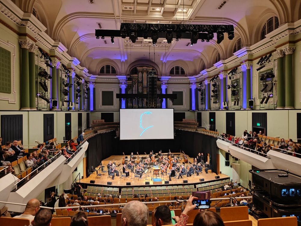 National Concert Hall Dublin with orchestra setting up on stage. A cinema screen has a sketch of Alfred Hitchcock's profile projected on to it