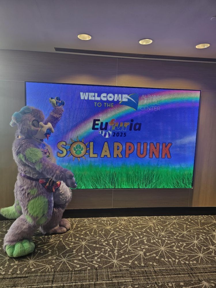Whimsy, a full digi fullsuit of a purple dog, poses in front of the sign for Eufuria 2025: Solarpunk