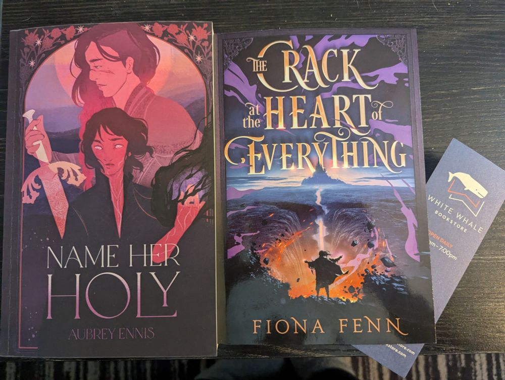 Side by side are books Name Her Holy and A Crack at the Heart of Everything. Also a bookmark for White Whale Bookstore.