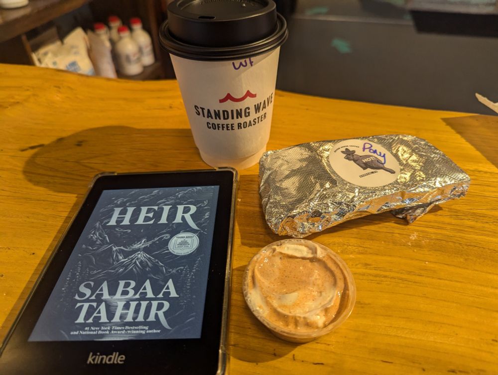 Table with: cover for Heir by Sabaa Tahir displayed on Kindle, cup of coffee from Standing Wave Coffee Roasters, a wrapped breakfast burrito, and sauce.