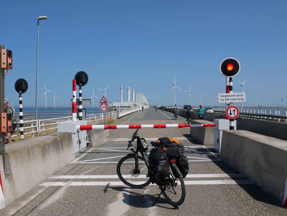 Das Foto wurde entlang des längsten Deiches der Delta-Werke in den Niederlanden aufgenommen. Im Vordergrund sieht man mein Trekkingrad mit großem Gepäck vor einem Schlagbaum, der den Autoverkehr von der Fahrradspur ausschließt mit einem rot leuchtenden Warnlicht. Dahinter beginnt der Deich, zu erkennen an den Hebewerken für die Fluttore. Weiter im Hintergrund ein ein paar Windkraftwerke zu sehen, darüber der blaube Himmel.
