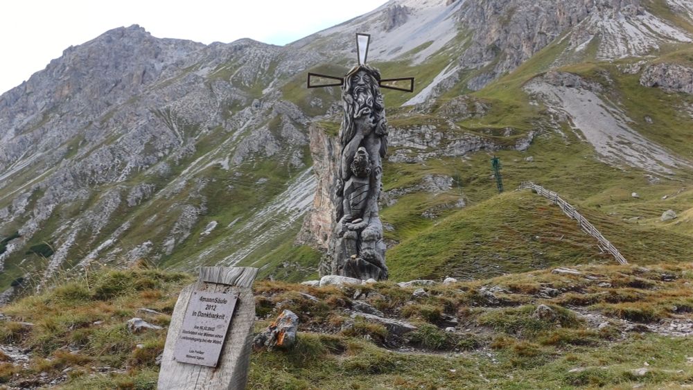 Das Bild zeigt ein etwas skuril anmutendes Denkmal in Form einer Säule aus Holz, aus der verschiedene Gestalten herausgearbeitet wurden, ähnlich einem Marterpfahl. Die obere Gestalt hat drei Strahlen vom Kopf ausgehend, könnte ein Heiligenschein sein, darunte sieht man einen weiteren Kopf, allerdings kopfüber, diverse Arme und Beine. Davor steht erklärend ein Schild 
"4mannSäule
2012
in Dankbarkeit

Am 06.02.20212
überlebten vier Männer einen Lawinenabgang bei der Wand.

Luis Pahitzer
Raimund Spiess"

Im Hintergrund beeindruckendes Alpenpanorama.