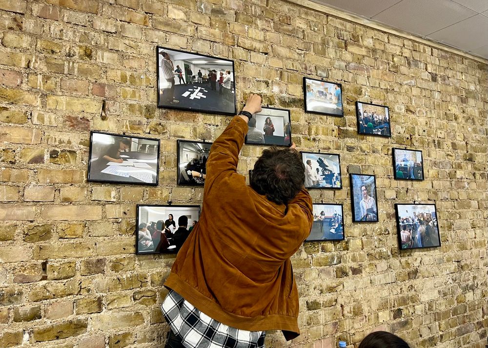 Man adjusting framed photo on yellow brick wall. Several photos with people writing and performing.