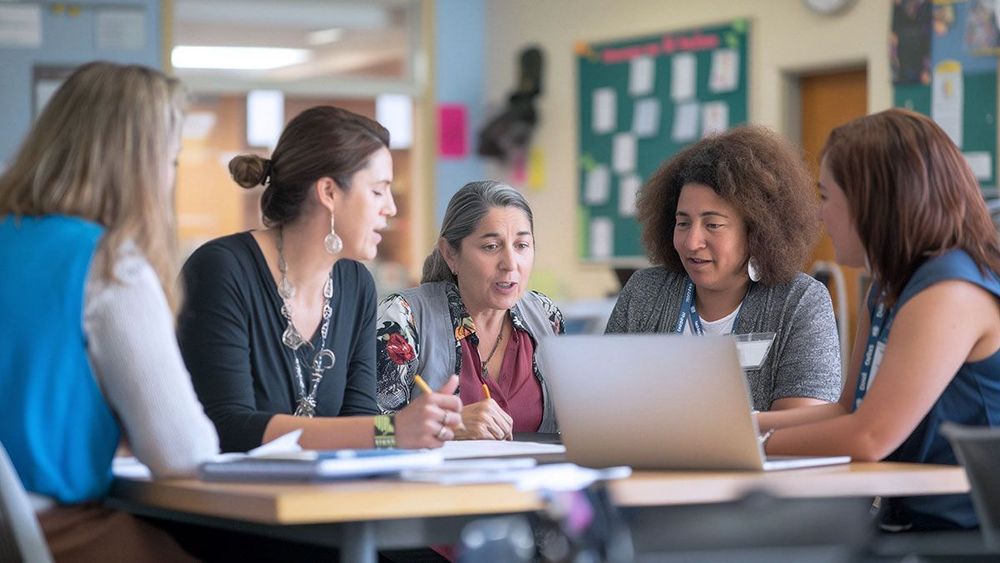 Five people collaborating at a table in a classroom setting, with books and a laptop on a desk