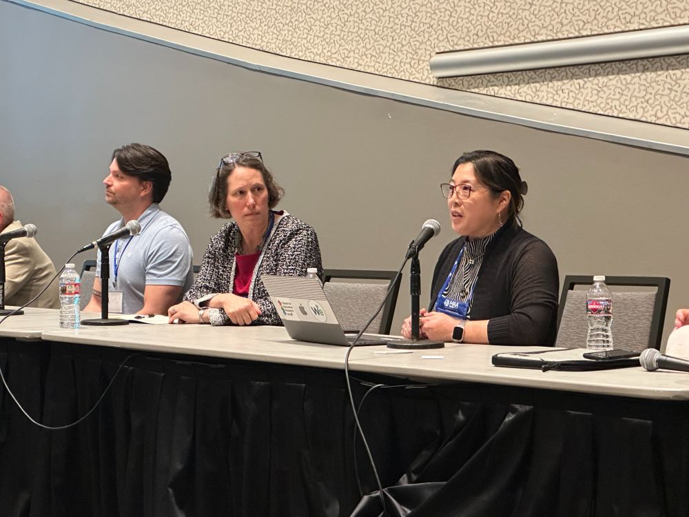 Four panelists, including one speaking, seated at a table with microphones and laptops during a conference session.
