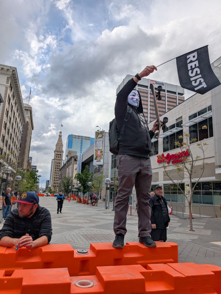 A man in a Guy Fawks mask waves a black flag that says resist