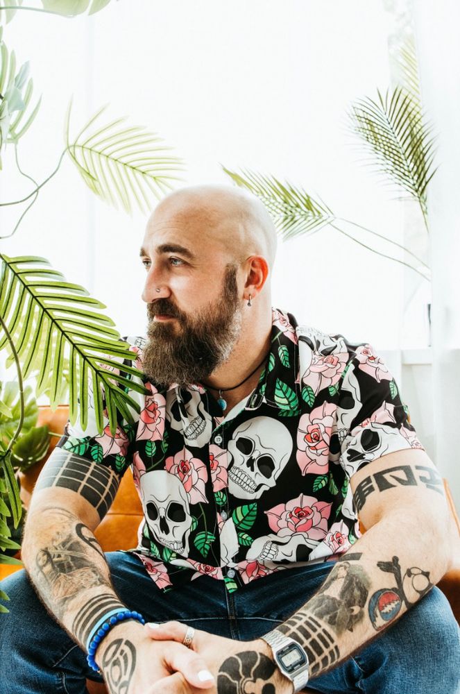 musician / producer Nmesh wearing a button up shirt with skulls and flowers on it. there are plants and leaves around him in a light background 