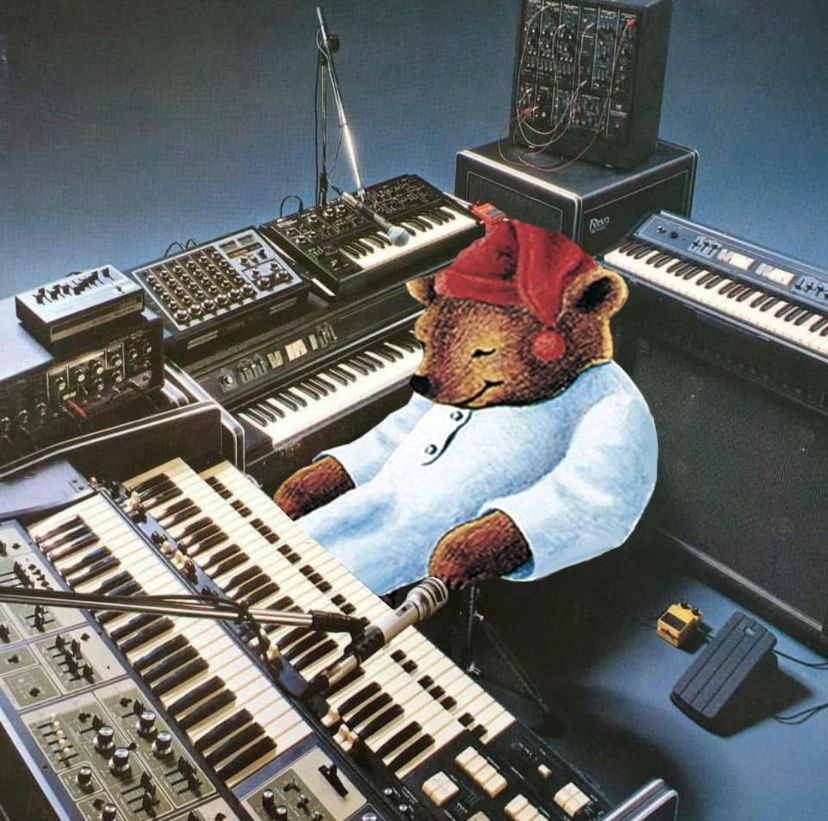 a bunch of synths / keyboards, effects pedals, microphones, a modular synth(?) and a bunch of other gear arranged around the sleepytime bear 