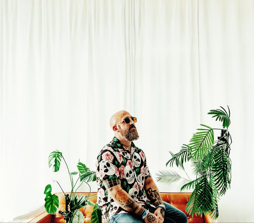photo of producer Nmesh wearing a shirt of skulls and flowers. he is sitting near an orange couch surrounded by plants with a large white curtain behind him. he is wearing aviator sunglasses 