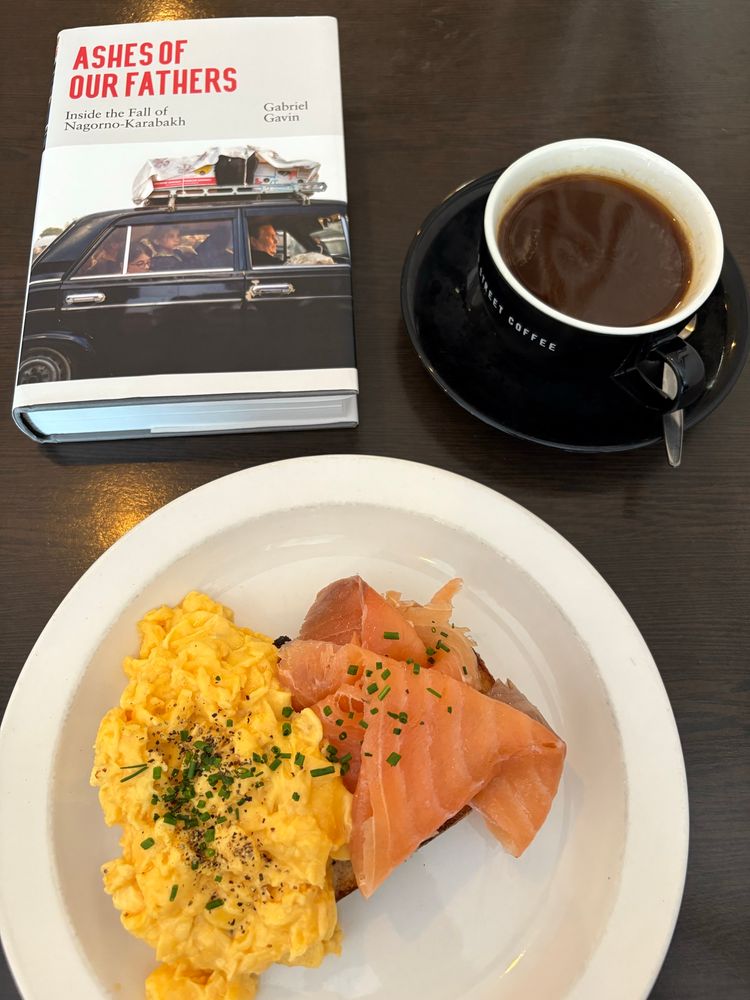 A plate of salmon and scrambled eggs on a dark table top. Just above it are a cup of black coffee and a copy of Ashes of Our Fathers by Gabriel Gavin