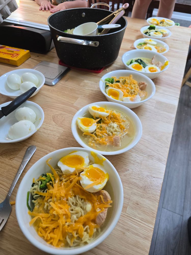 Seven white bowls of ramen with chicken, soft boiled eggs, wilted spinach and broccoli, and cheddar cheese sit on a wooden countertop next to a couple bowls of peeled eggs and a pot of noodles.