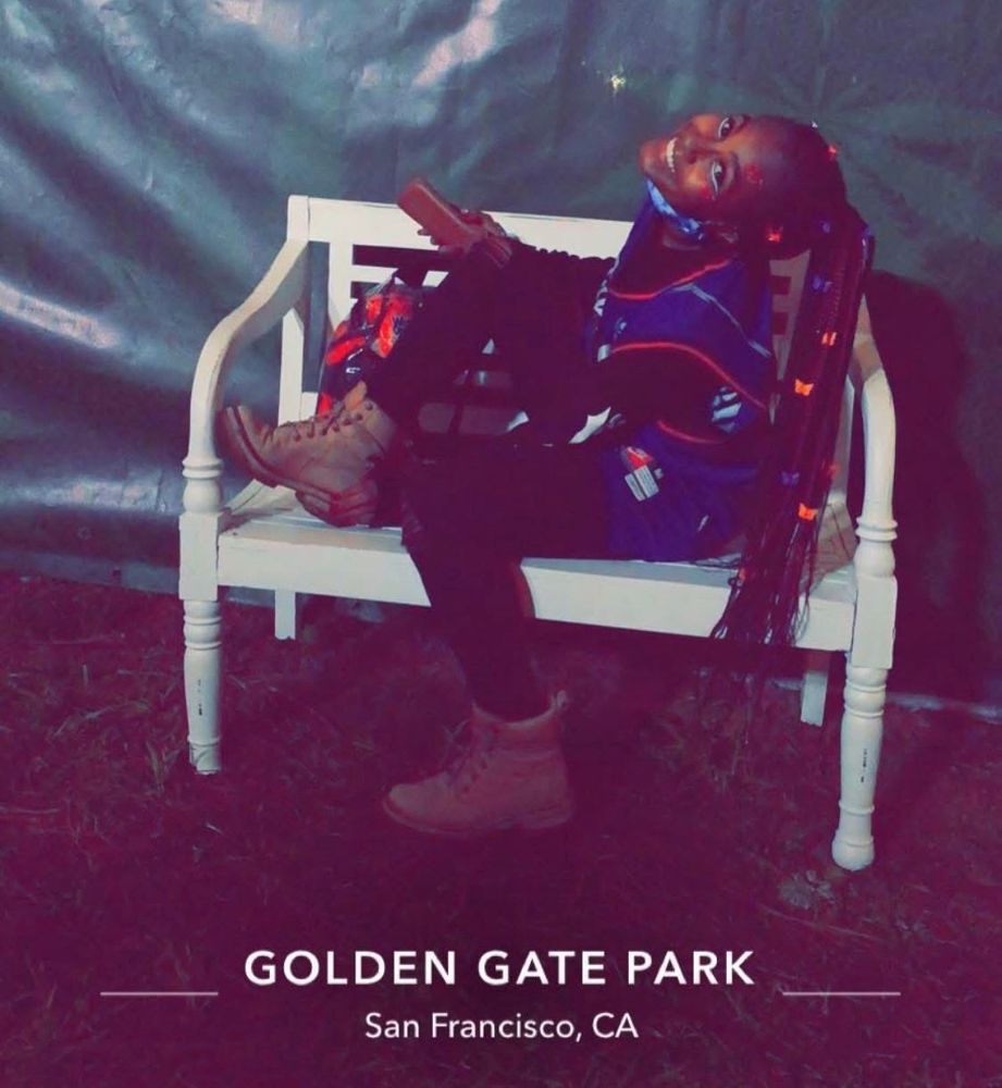 Beautiful black woman is smiling in a braided ponytail wearing a Vince Carter, basketball jersey, black jeans and dirty timberlands while sitting on a white park bench at Golden Gate Park in San Francisco, California