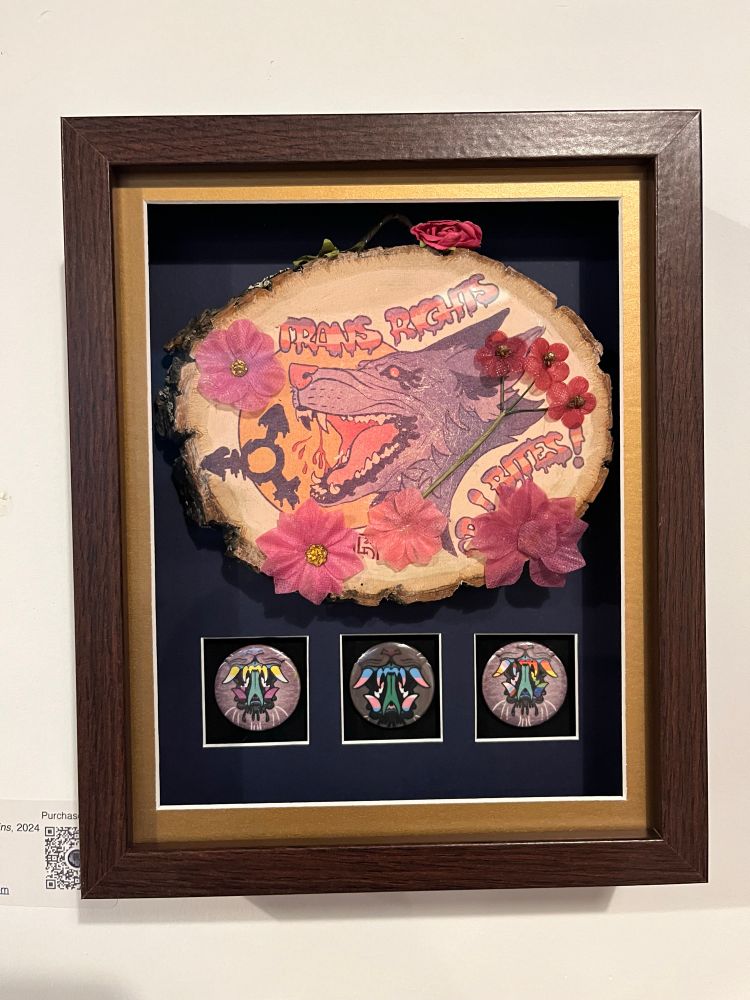 a brown shadow box frame with a wooden plaque inside that has a snarling wolf and the caption “trans rights or i bites!” with three pins underneath that have monster teeth colored as the nonbinary, trans, and progress flags respectively. it’s matted in blue with a small gold border against the glass.