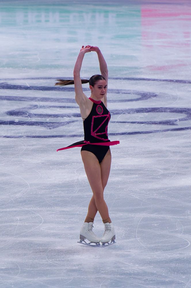 Lara Naki Gutmann Women's short program skating in a black with pink trim outfit with the Squid Games shapes on the back.  Performing a spin