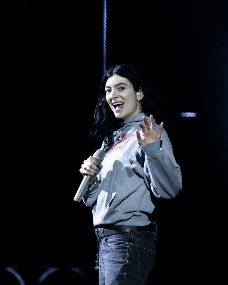 Lorde on stage in Seattle. Grey hoodie and jeans, waving, big smile. Ultrasound World Tour.