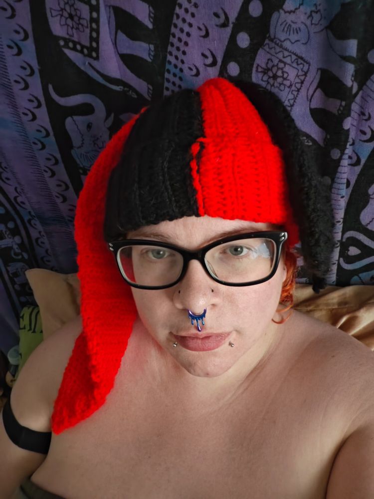 White femme presenting person wearing glasses and a crochet split red and black beanie with opposing coloured rabbit ears but the black ear is finished half way to look like it was bitten off.
