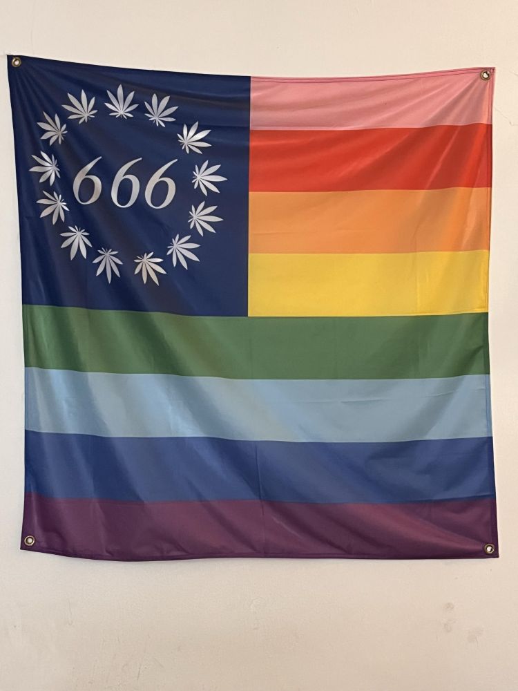 A Pride flag with the numbers 666 surrounded by a circle of cannabis leaves in the upper left corner, like the original stars on the US flag 