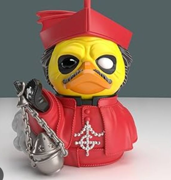 Foto de um pato de borracha caracterizado como o personagem Cardinal Copia, da banda Ghost. Ele usa uma roupa vermelha de cardeal com o símbolo da banda, chamado de grucifixo, em strass, e segura um incensário.