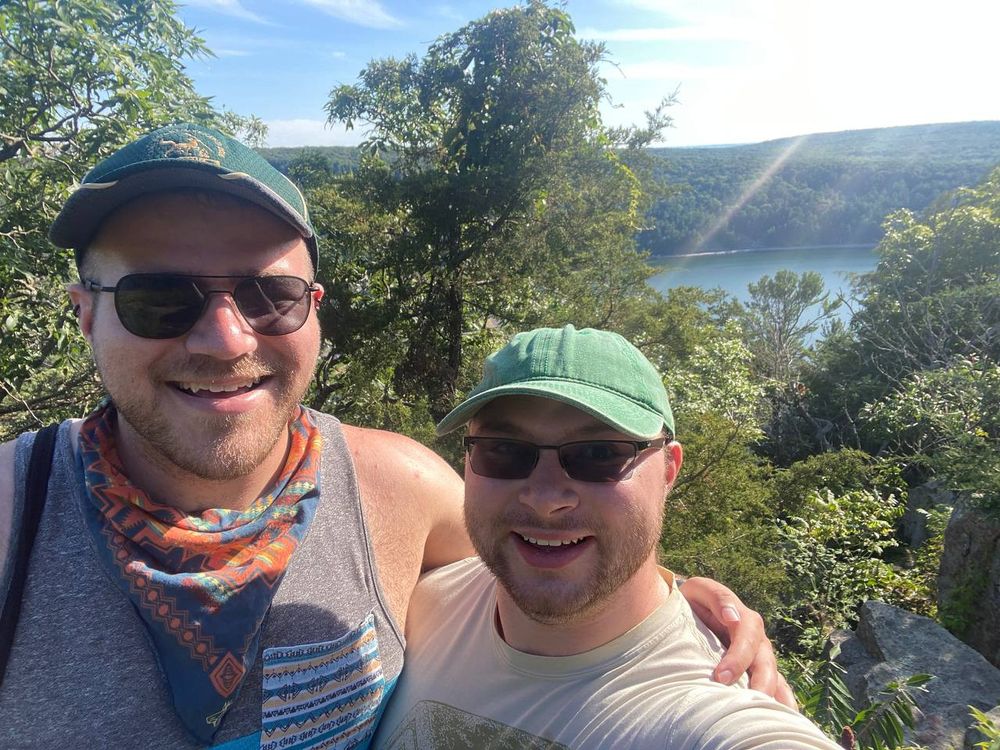 Two people stand in the foreground, wearing green hats and sunglasses. In the distance, a lake can be seen, with a geologic structure that holds the water in place visible above it, and the sun brightly obscuring the sky above that. Trees hold the background behind the people.